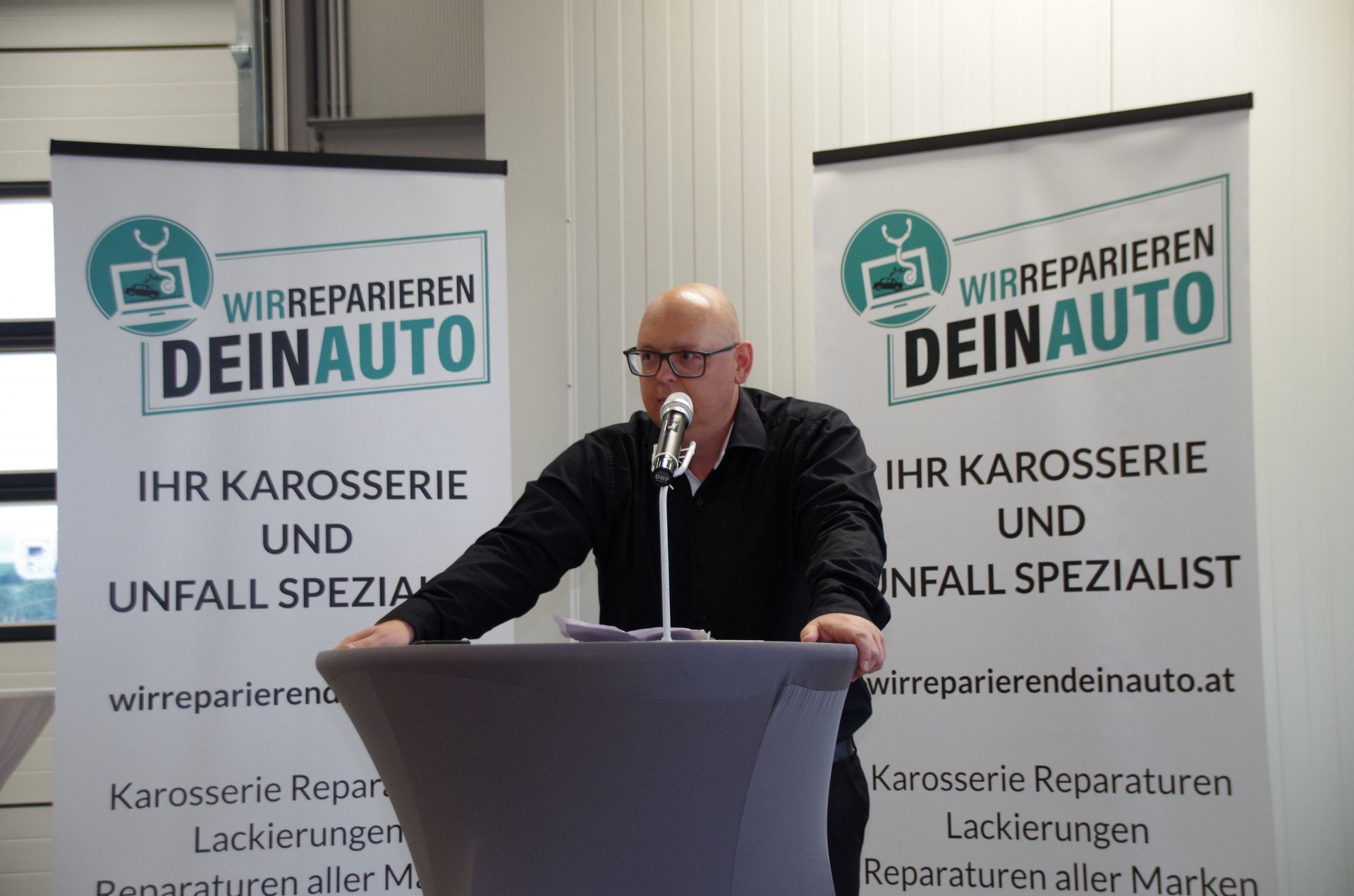 Man speaking at a podium before two banners reading “Ihr Karosserie und Unfall Spezialist”
