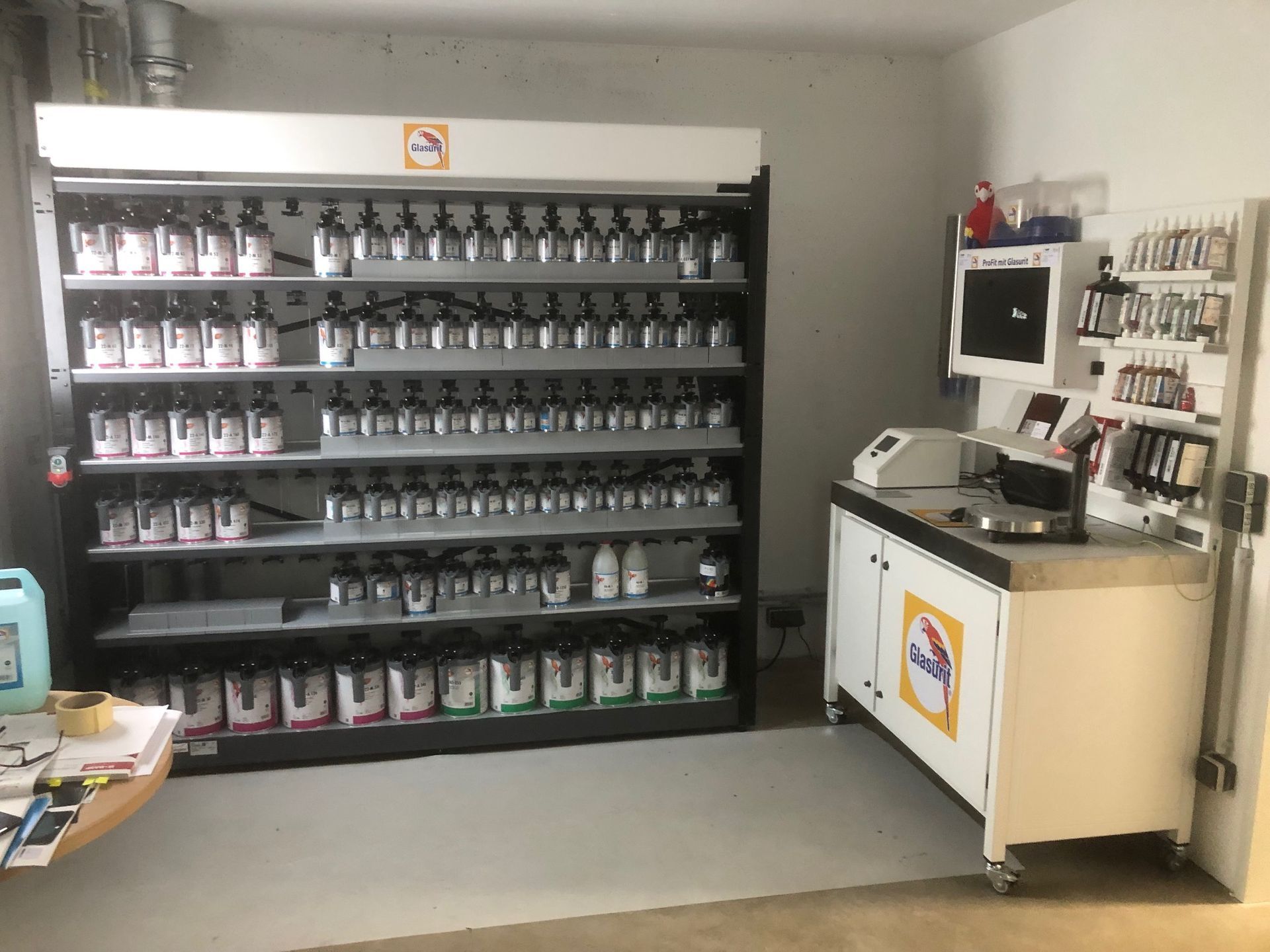 Lagerraum mit Regalen voller abgefüllter Produkte und einer Kasse vor einer grauen Wand.