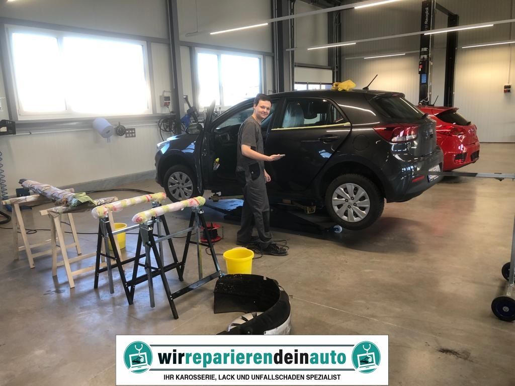 Ein Mann steht in einer Autowerkstatt neben einem schwarzen SUV. Werkzeuge und Eimer liegen auf dem Boden