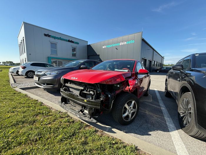 Beschädigter roter SUV mit fehlender Frontstoßstange und verbeulter Motorhaube vor einem Autohaus geparkt.