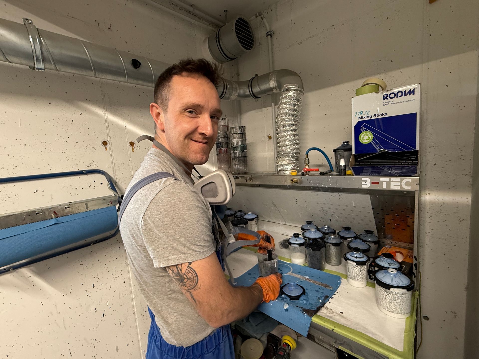 Mann arbeitet mit Farbspritzgerät in einer Werkstatt neben Lüftungskanälen und Farbdosen.