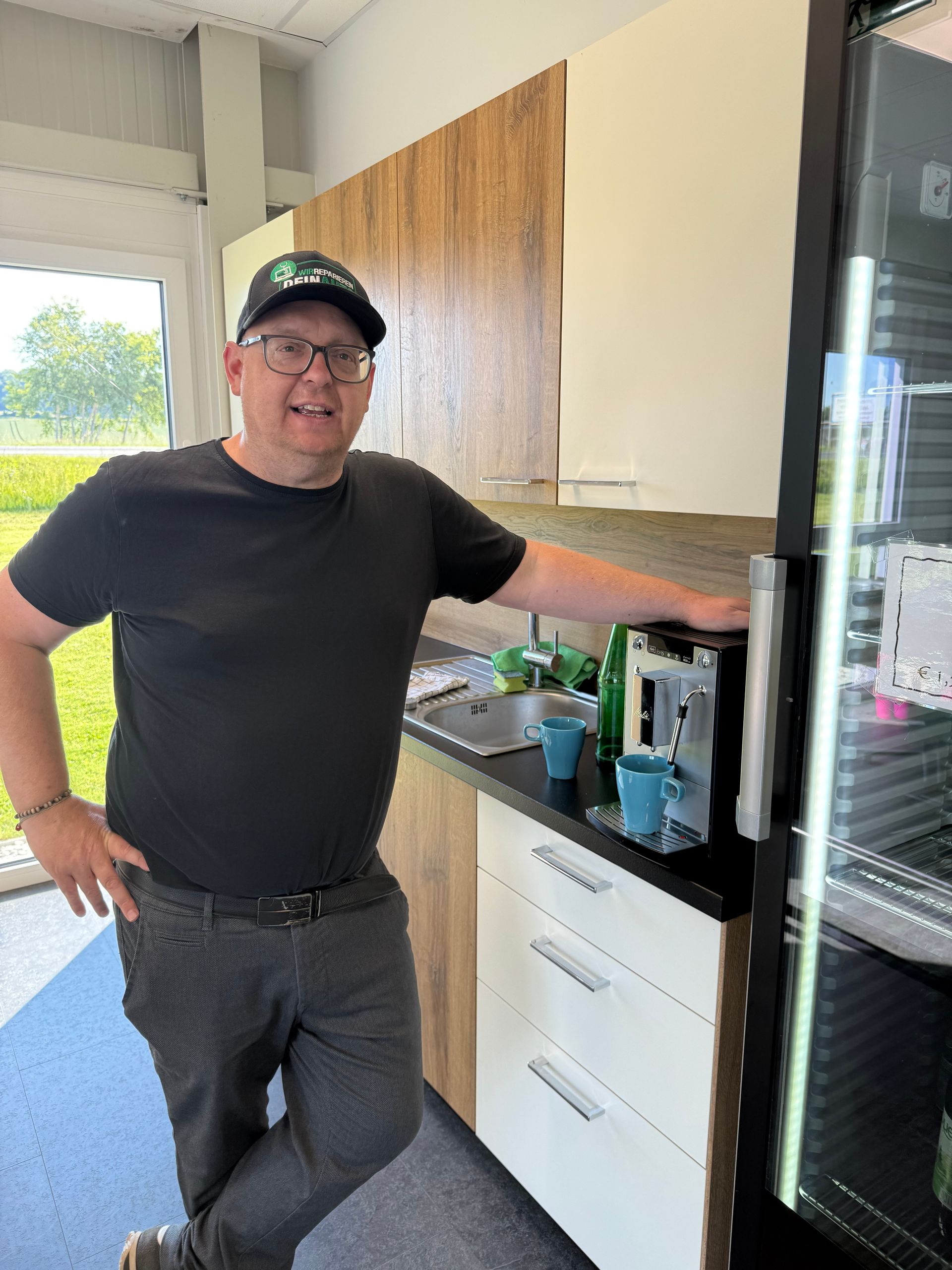 Ein Mann steht in einer Küche und lehnt sich an die Küchentheke neben einer Kaffeemaschine und weißen Schränken.