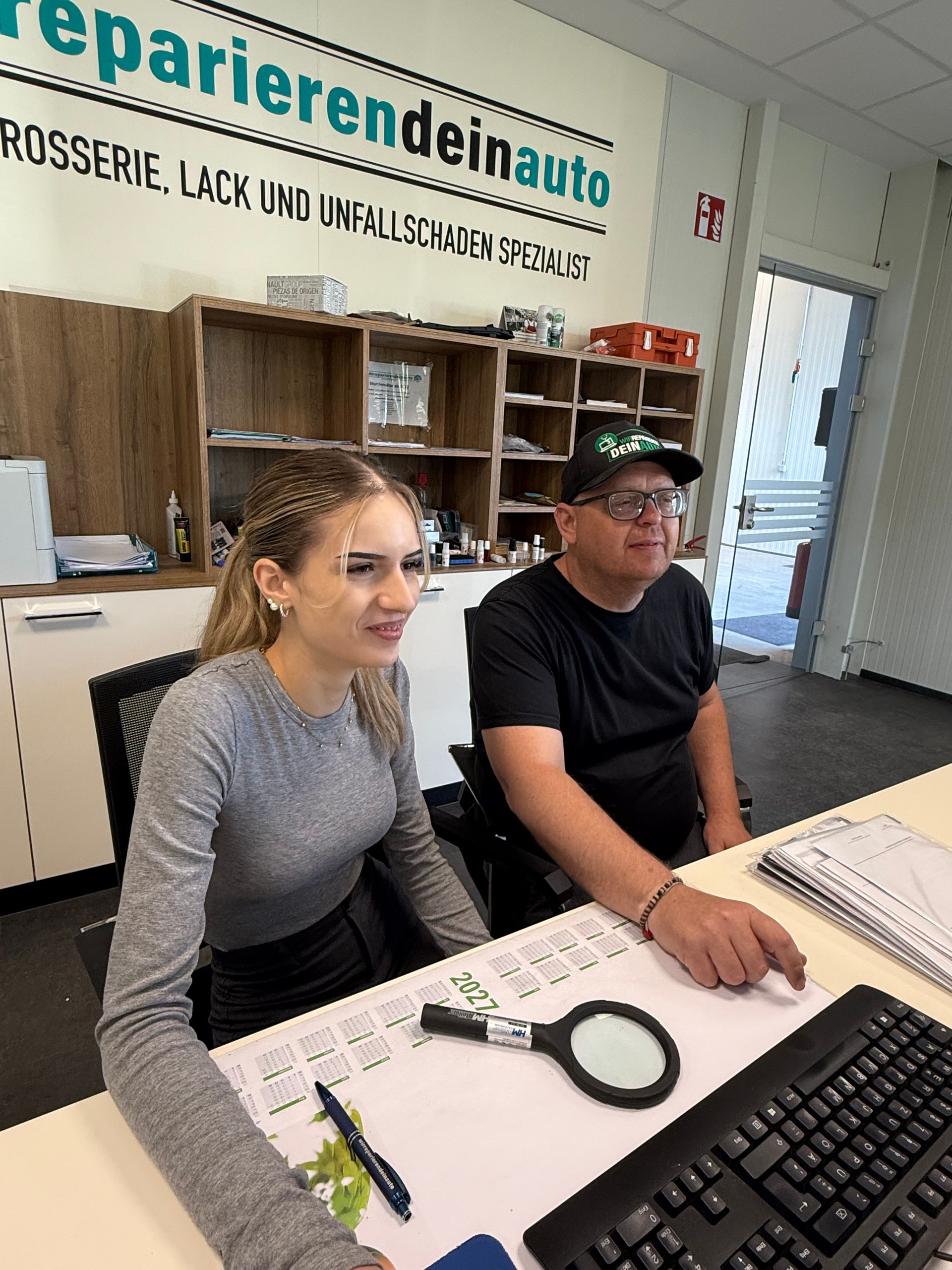 Zwei Personen an einem Schreibtisch im Büro einer Reparaturwerkstatt, im Vordergrund eine Computertastatur und Werkzeuge.
