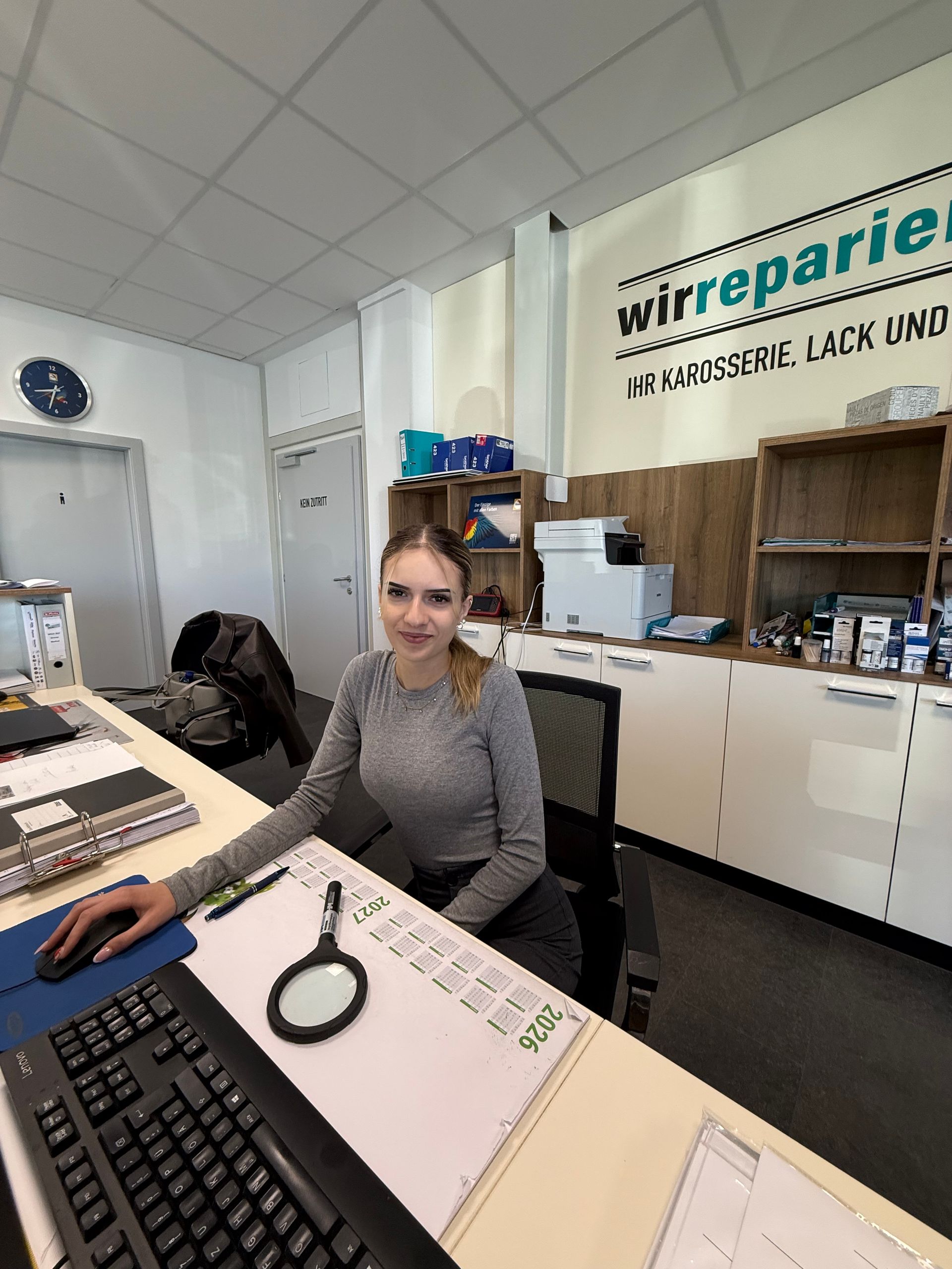 Eine Frau sitzt lächelnd an einem Schreibtisch im Büro einer Reparaturwerkstatt, neben einem Computer und Papierkram.