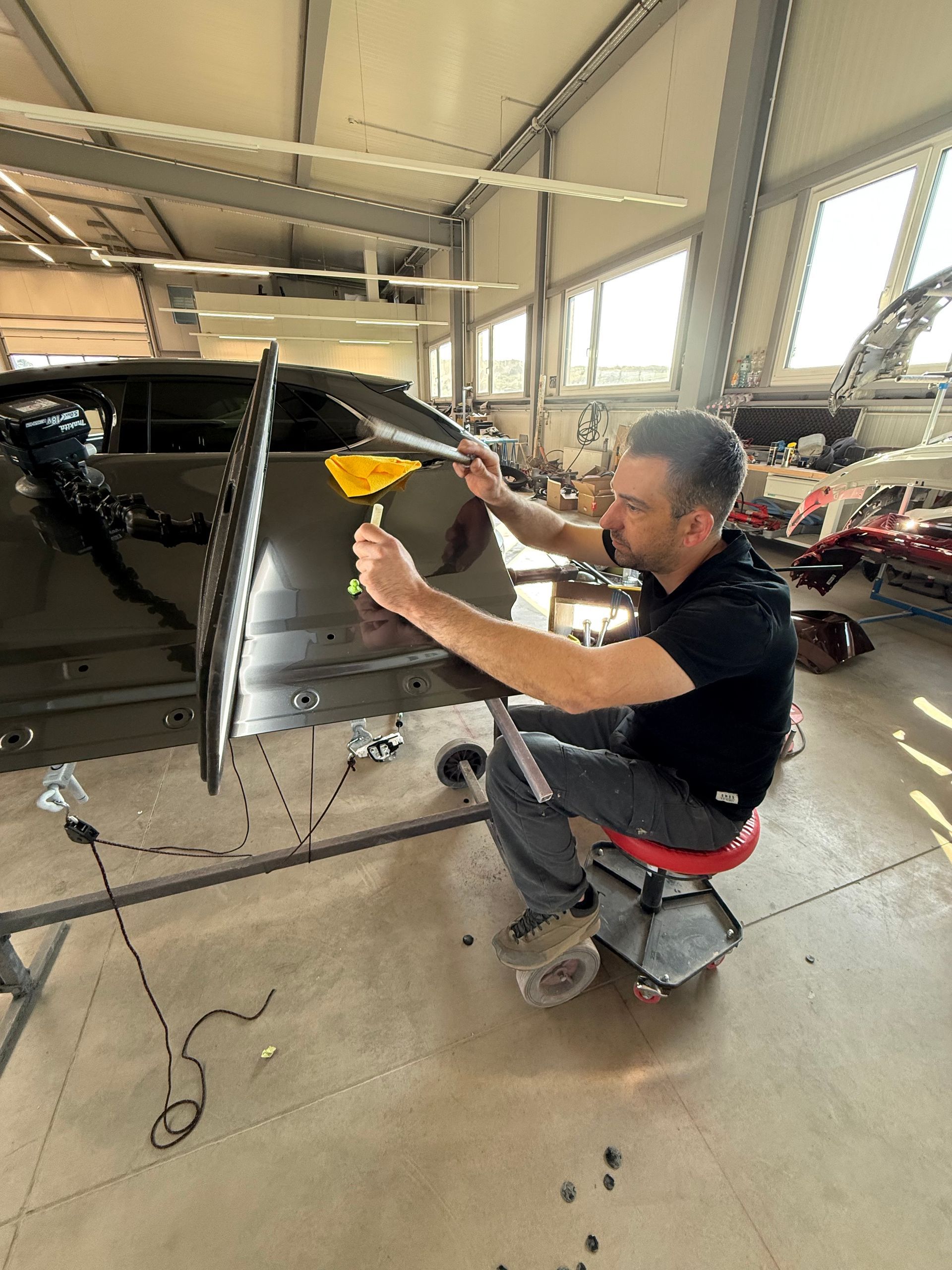 Ein Techniker poliert in einer Garage mit einem gelben Tuch in der Nähe eines Fensters ein schwarzes Auto.