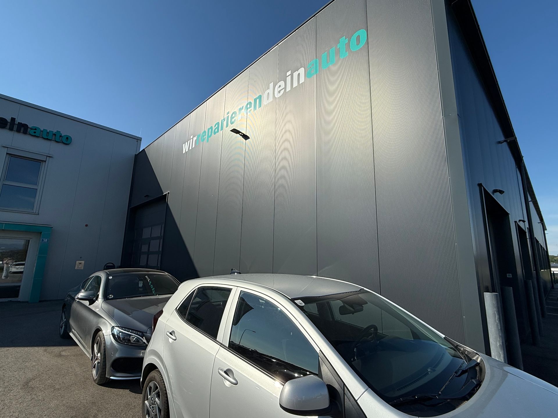 Zwei geparkte Autos neben einem grauen Gebäude mit türkisfarbener Aufschrift im hellen Sonnenlicht