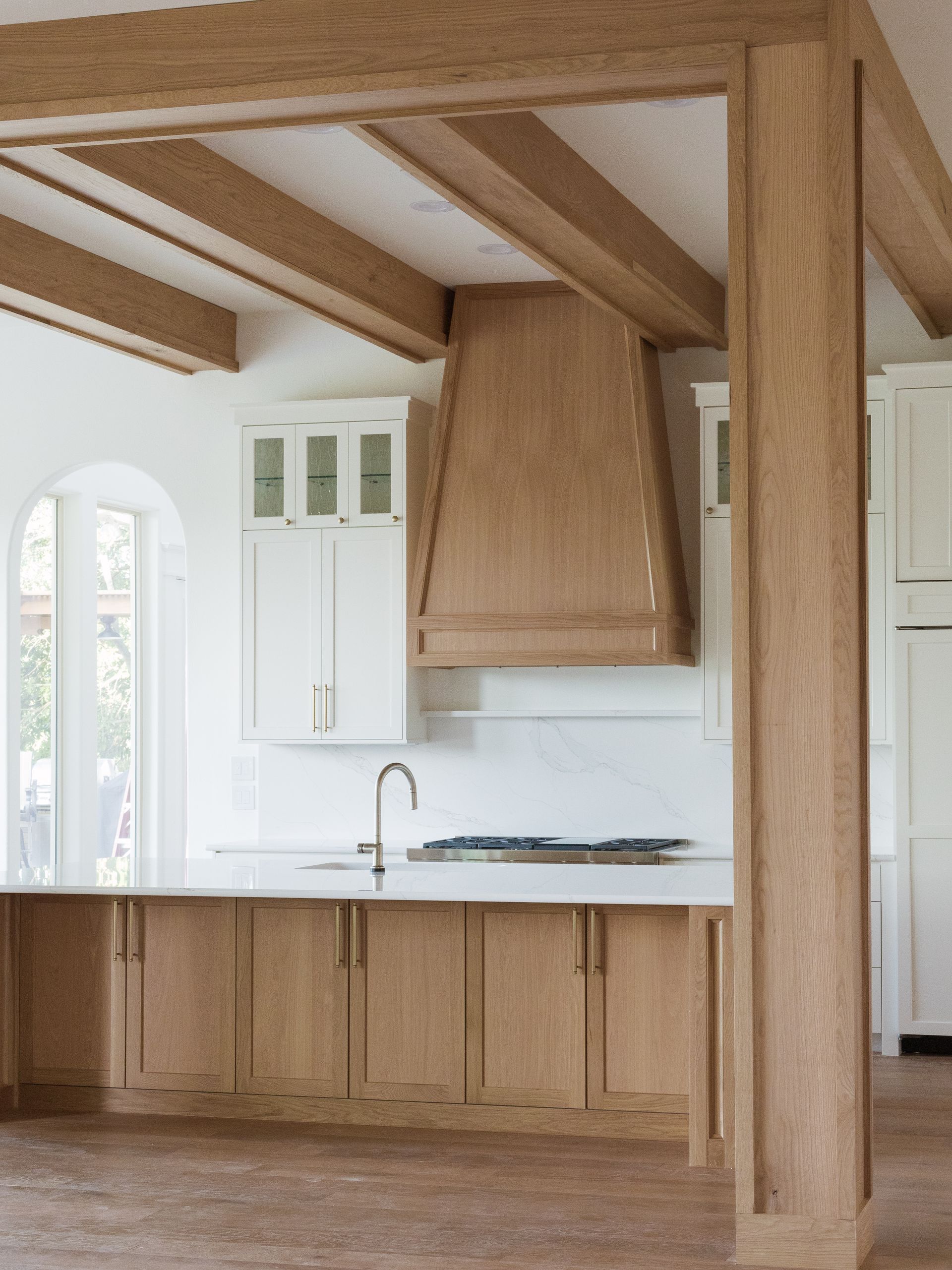 Modern kitchen with light wood beams, cabinets, and range hood. White countertops and walls.