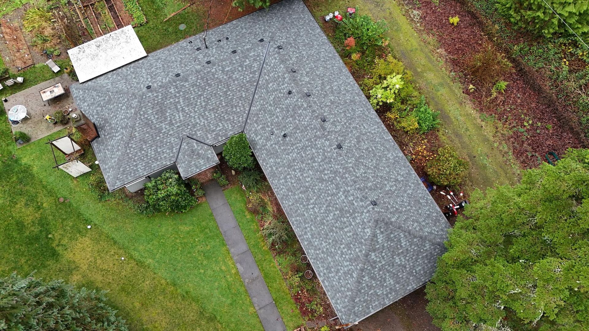 Overhead view of a dark shingle roof in an L-shape, surrounded by a yard, gardens, and trees.