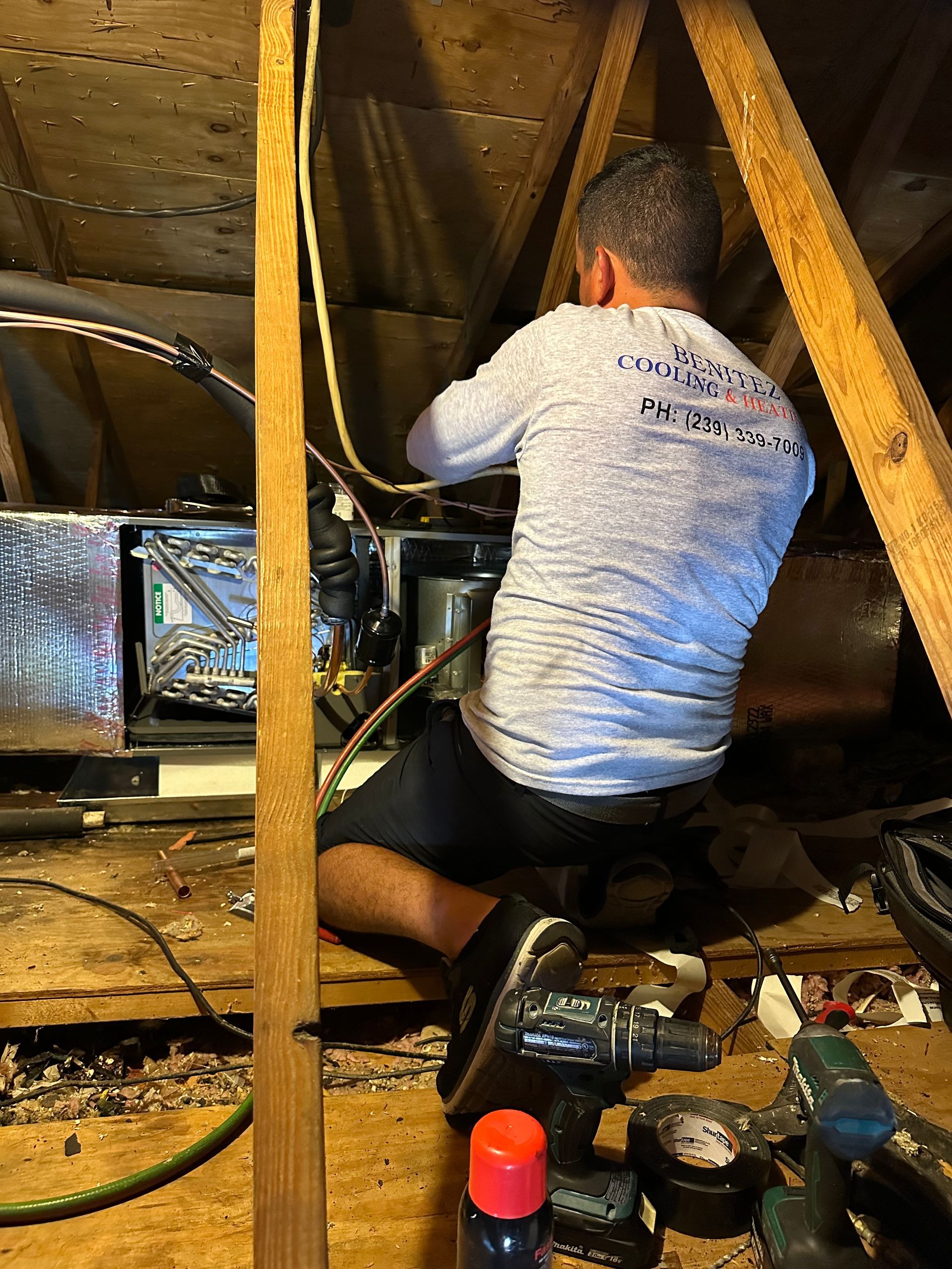 A man is kneeling down in an attic working on an air conditioner.
