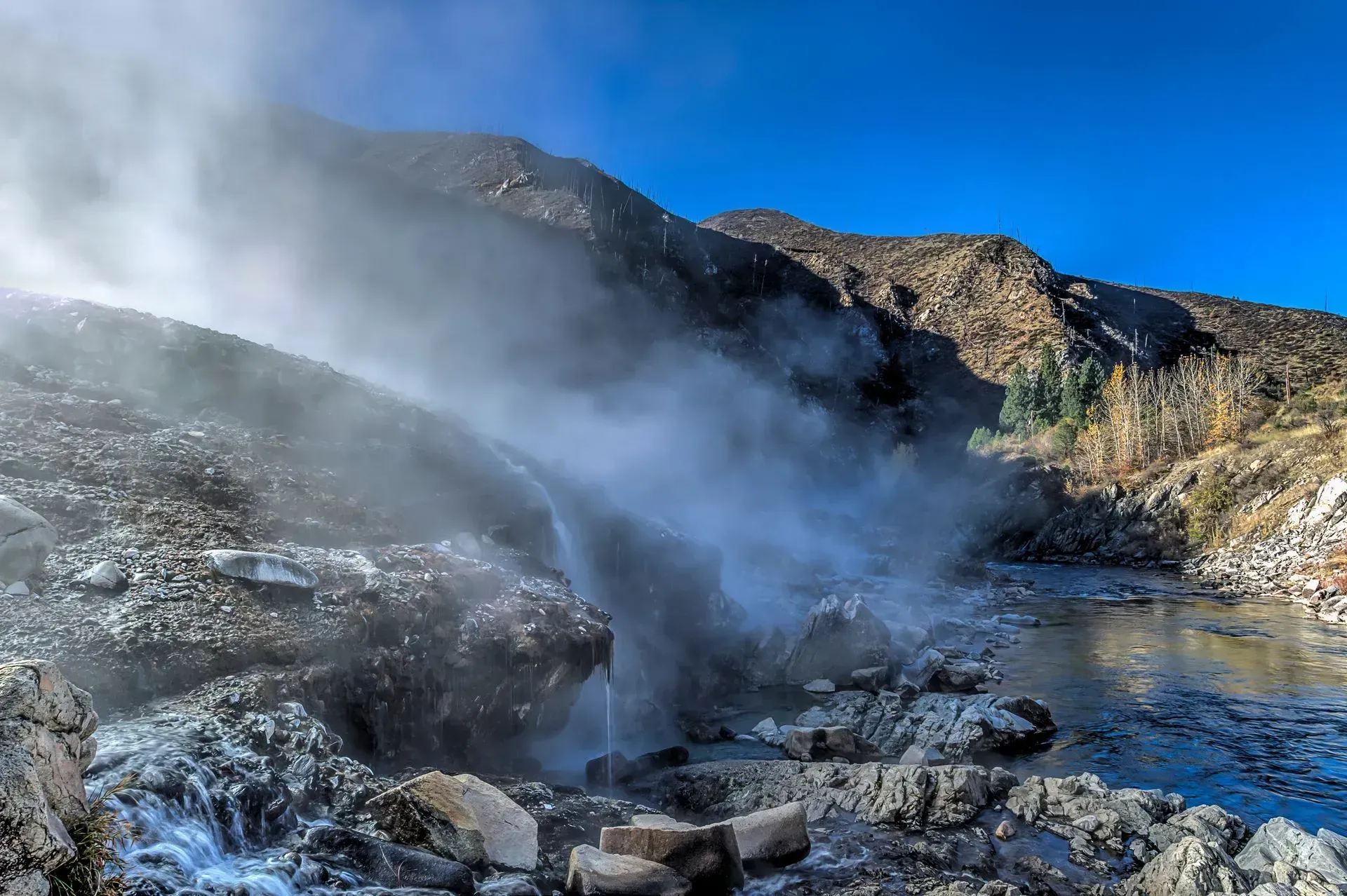 Steam rising from a hot spring cascading down a rocky mountainside on a sunny day.
