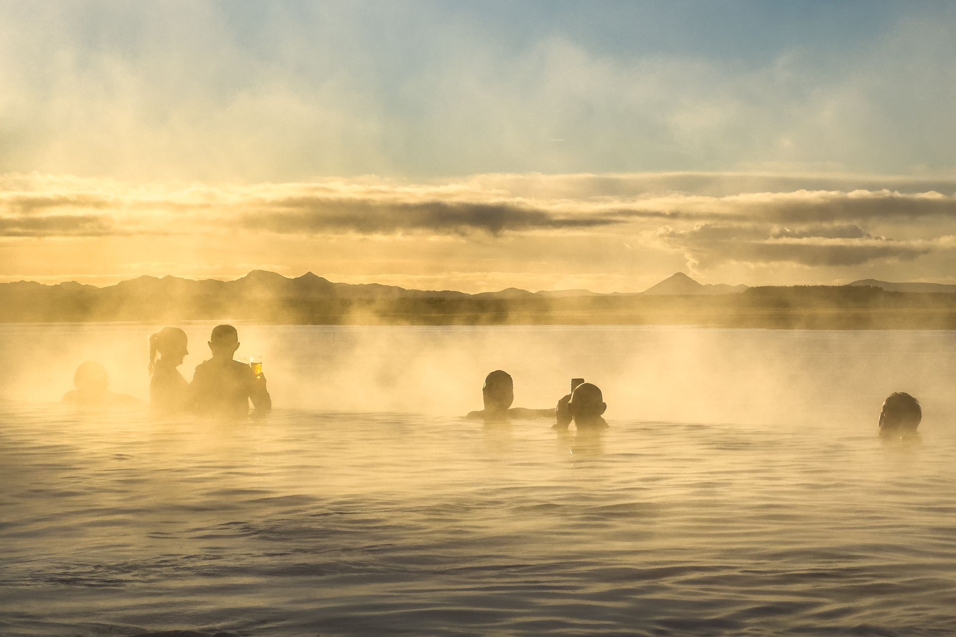 People bathing in a steaming hot spring at sunrise, with golden mist over calm water.