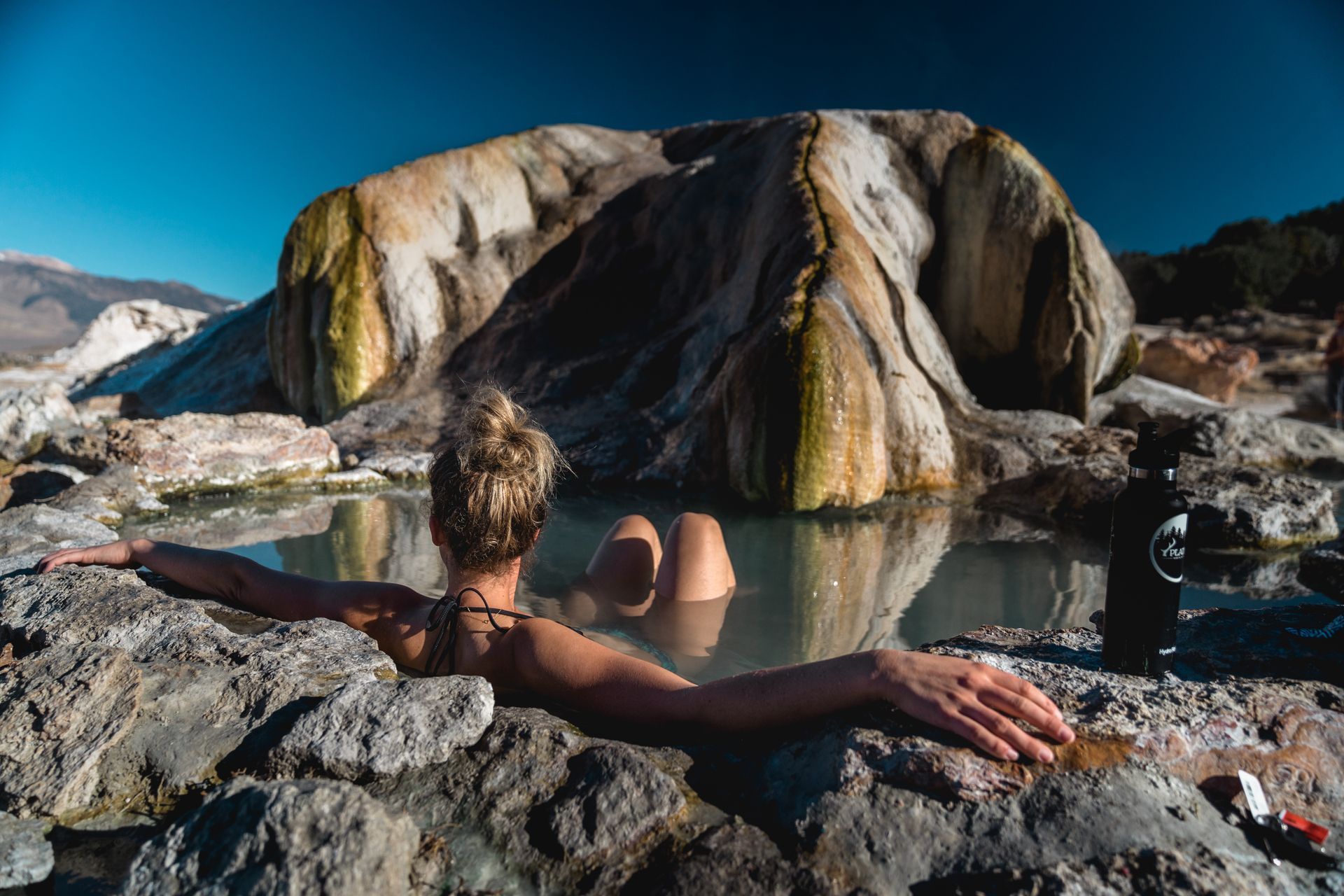 Person relaxing in a hot spring between large tan rock formations under a clear blue sky