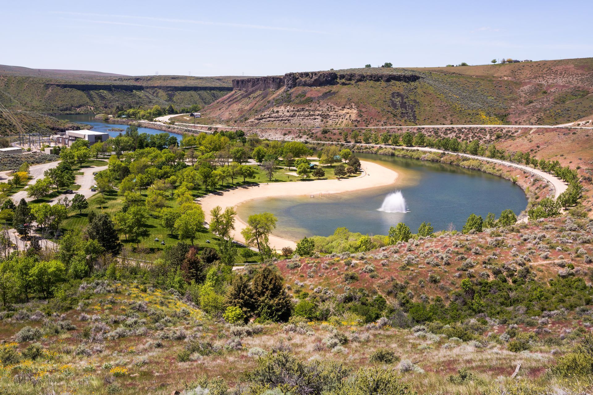 Scenic view of a lake with fountain, beach, and surrounding trees and brown cliffs.