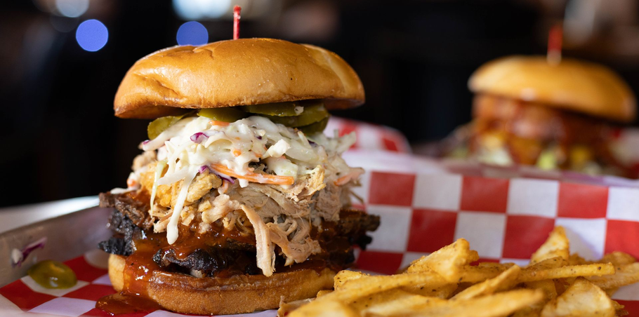 A piled-high burger with coleslaw, pickles, and pulled pork on a bun, with fries and another burger in the background.