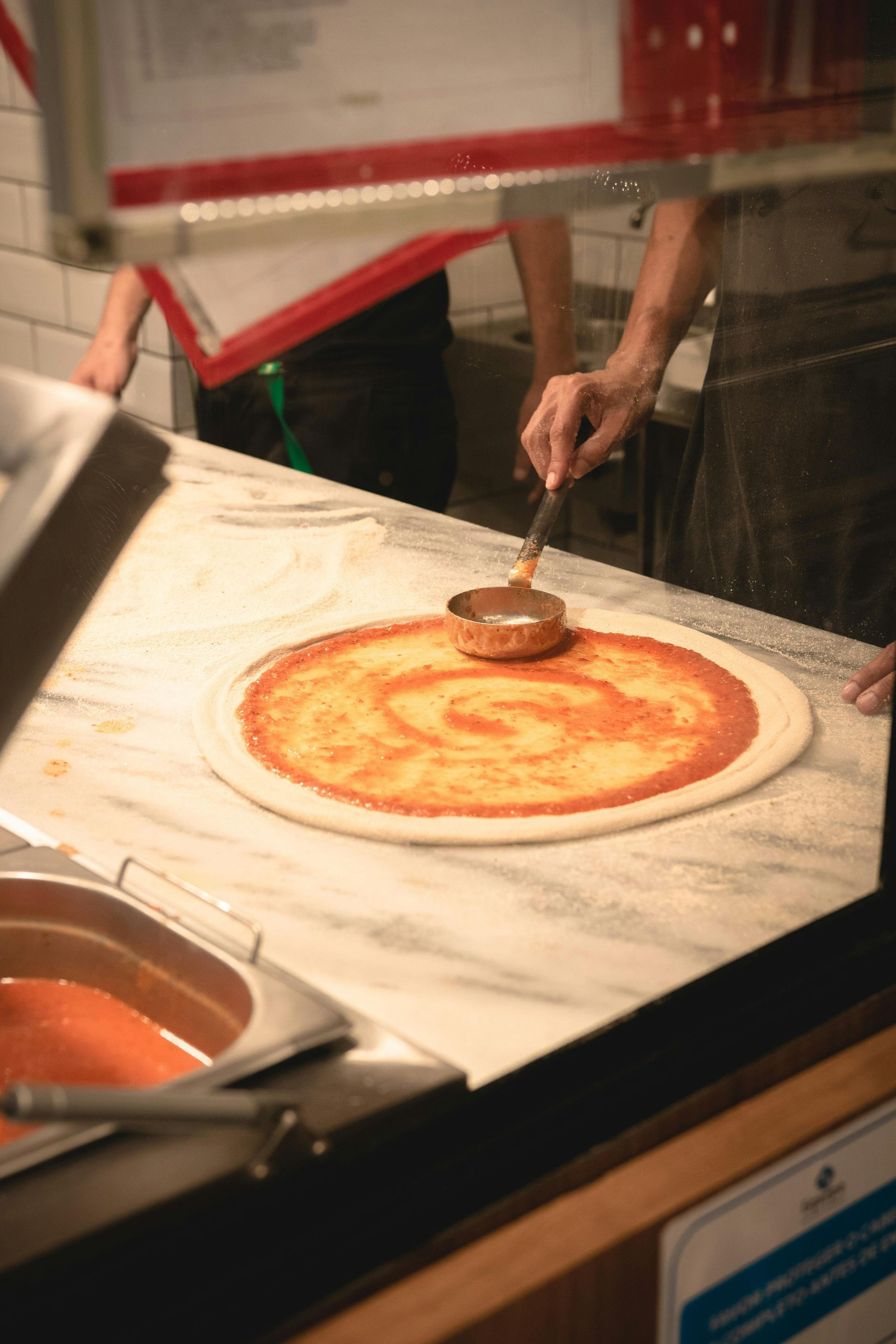 Person applying tomato sauce to pizza dough with a ladle on a marble counter.