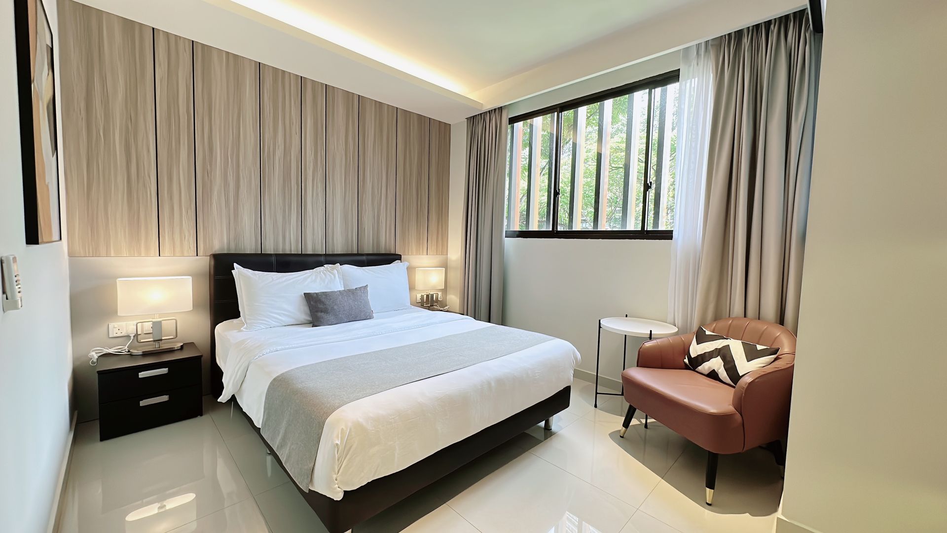 Modern bedroom with a neatly made bed, bedside table, armchair, and large window with curtains.