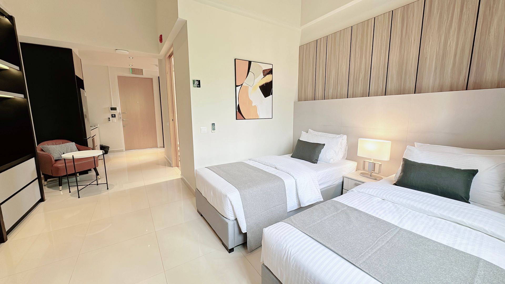 Bright hotel room with two single beds, beige decor, a lamp, and an open doorway to the hall