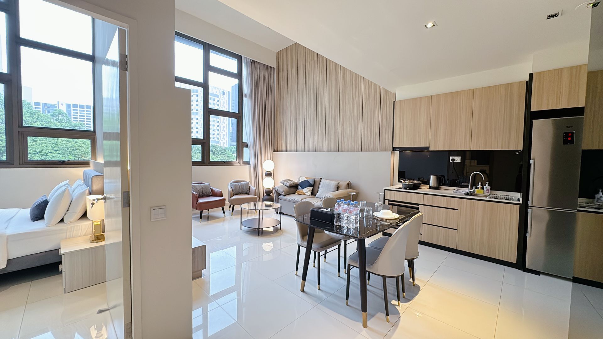 Modern open-plan living room and kitchen with dining table, light wood cabinets, large windows, and glossy white floors