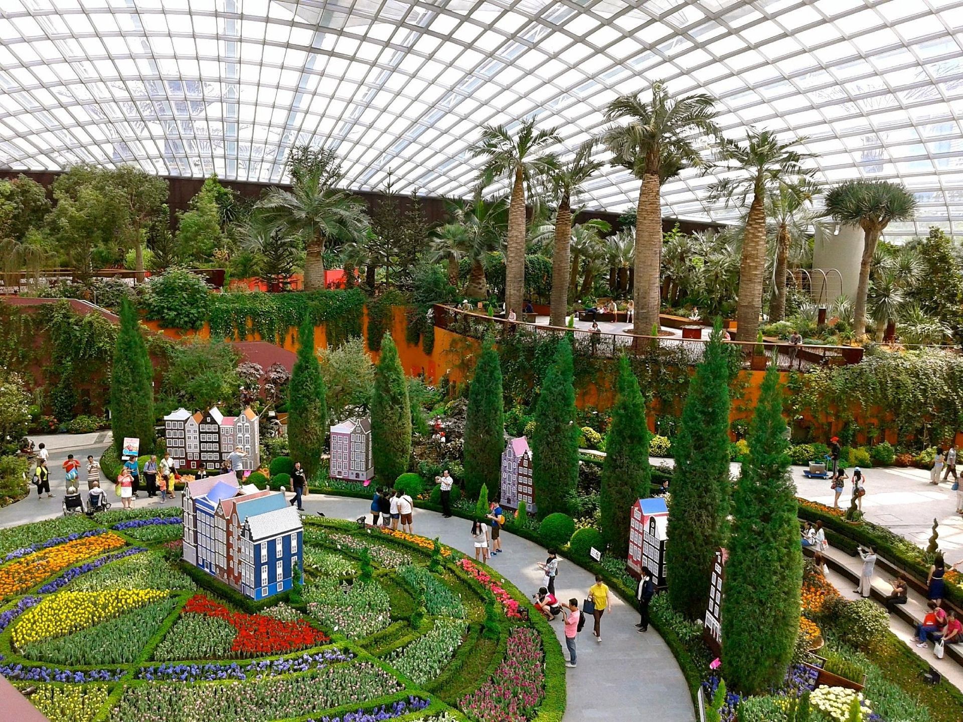 Indoor garden with colorful flower beds, tall palms, and people walking under a glass dome