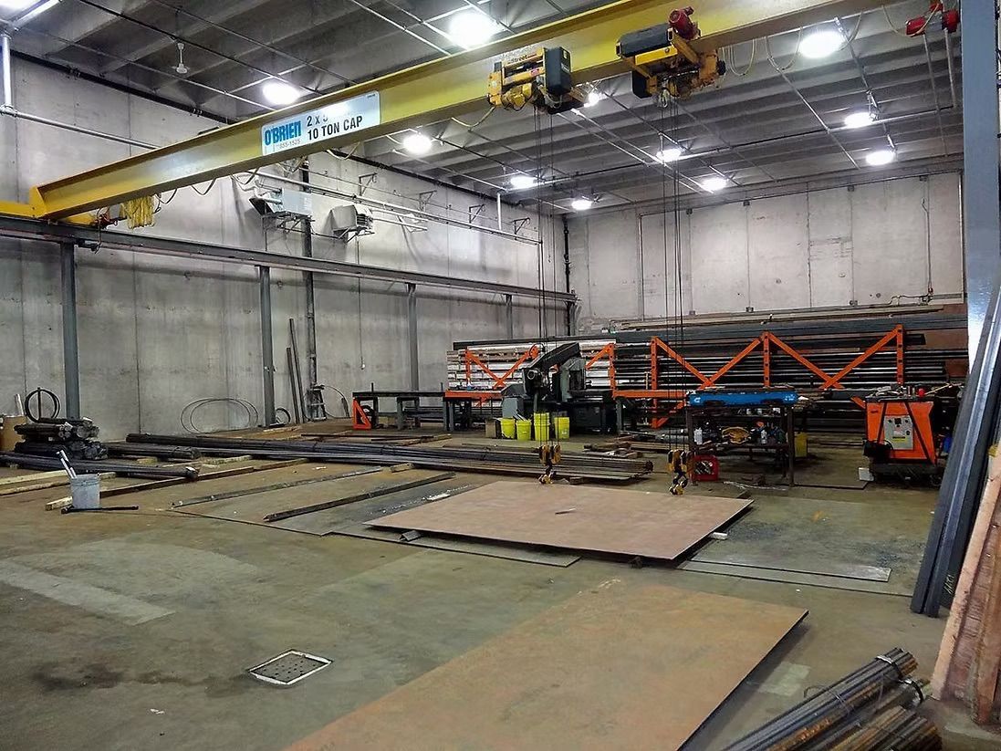 Industrial warehouse interior with a yellow overhead crane, metal shelving, various steel sheets, and floor equipment.
