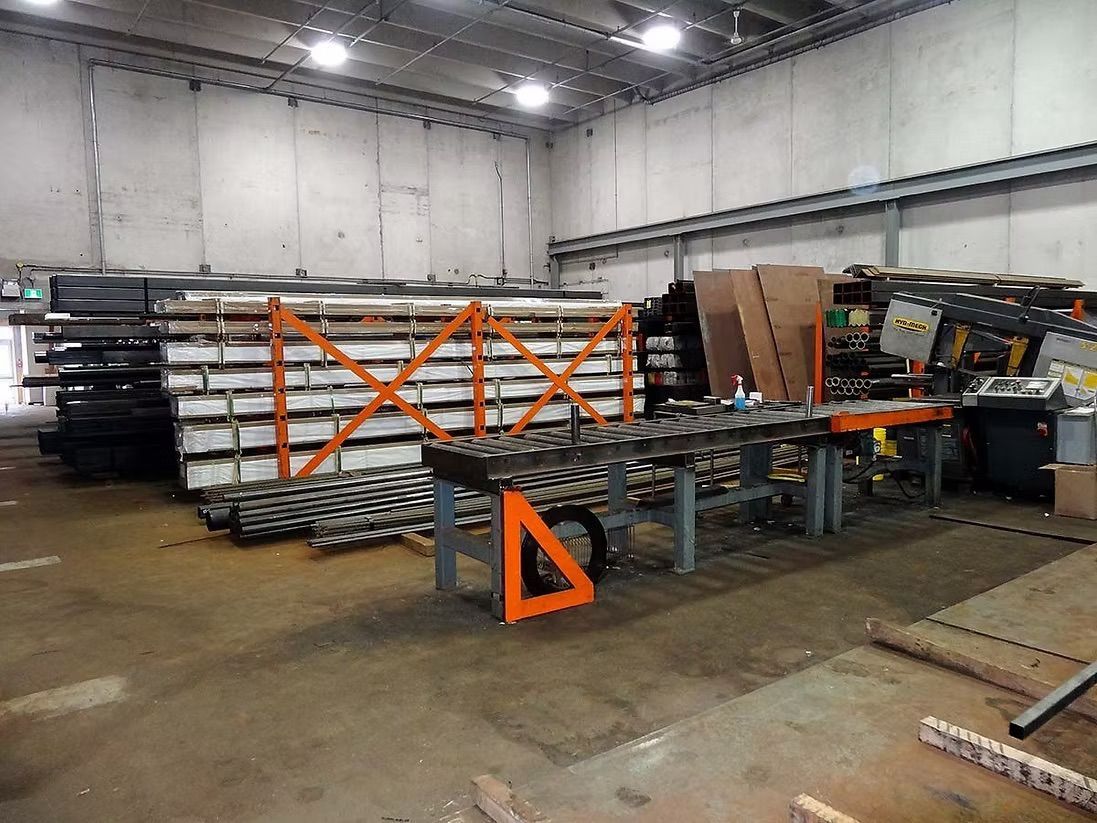 An industrial warehouse floor featuring stacks of metal beams on orange racks and a roller conveyor table.
