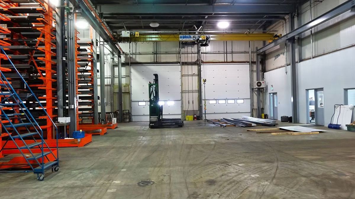 A wide, industrial warehouse interior featuring metal storage racks, a forklift, overhead crane, and loading bay doors.