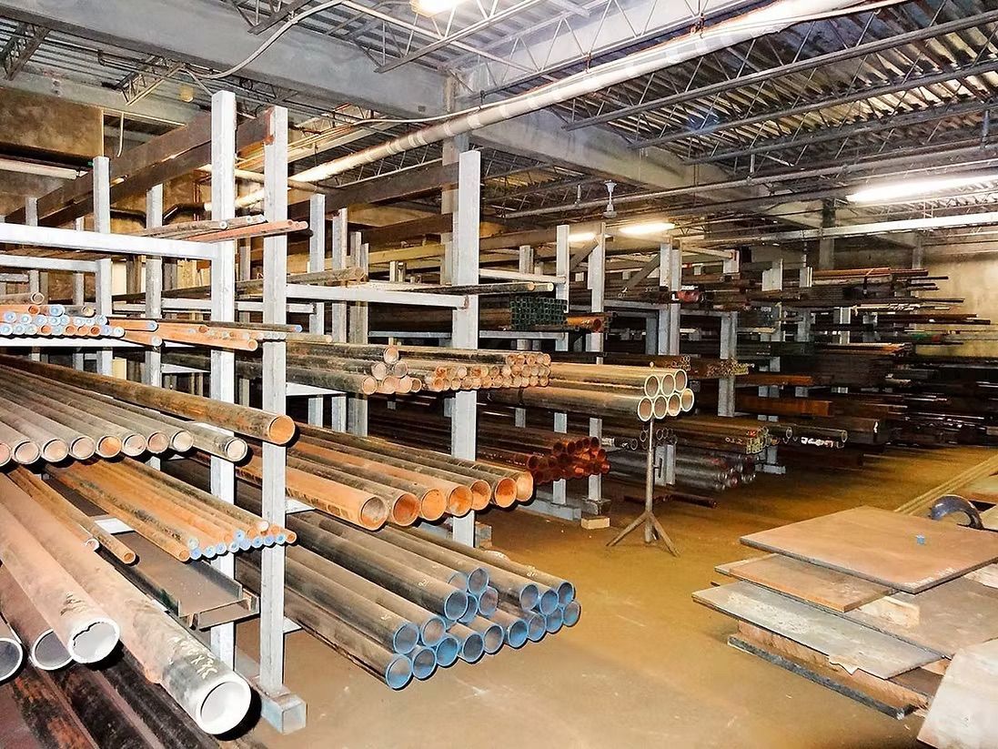 A warehouse interior featuring metal pipes, tubes, and flat sheets organized on industrial storage shelving racks.