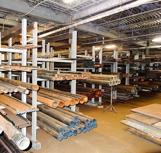 A well-lit warehouse interior featuring rows of metal pipes and flat steel sheets stored on industrial shelving racks.