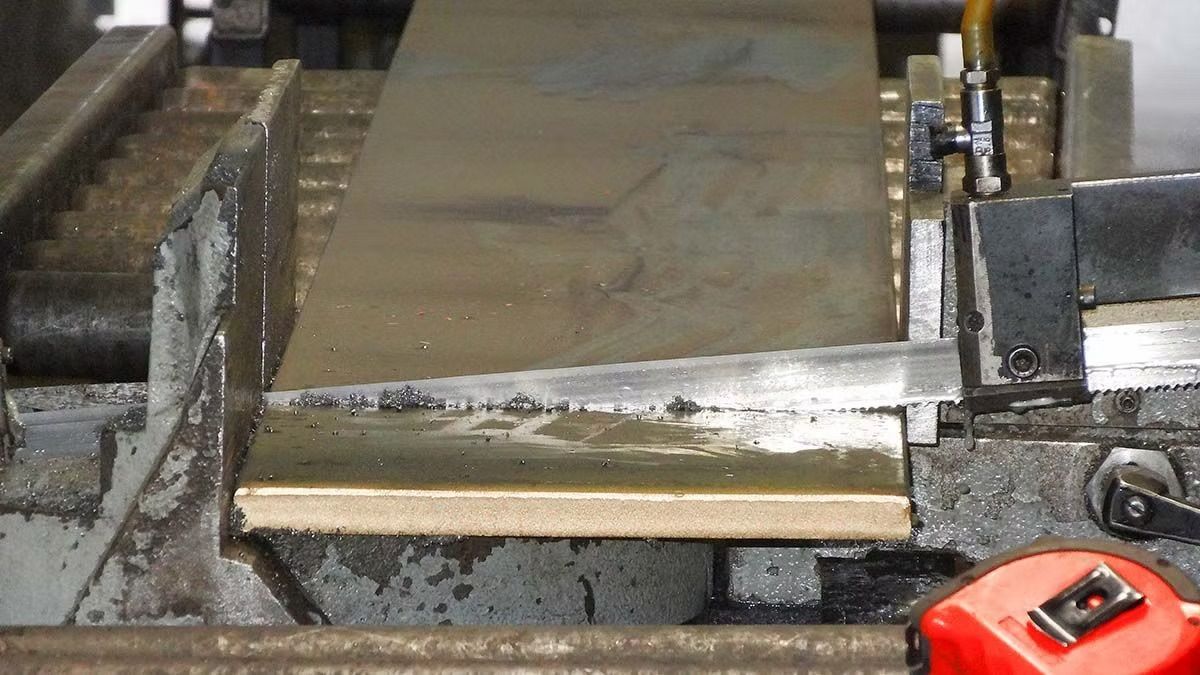 A horizontal band saw cutting through a thick metal plate held in a heavy-duty industrial vise.