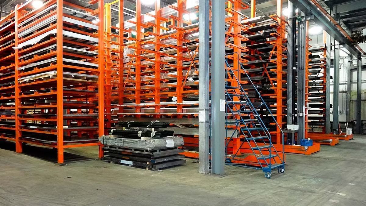Rows of orange metal industrial storage racks filled with sheets and bars of material in a warehouse setting.
