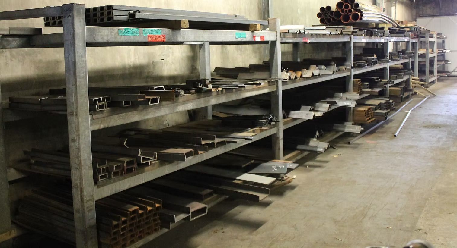 Metal industrial shelving in a workshop filled with various steel beams, pipes, and flat metal stock.