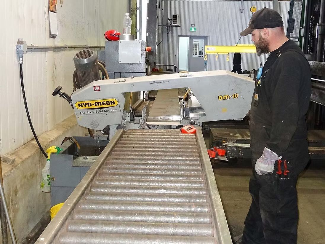 A worker in dark coveralls and a cap stands by a grey industrial band saw in a workshop with a roller conveyor system.