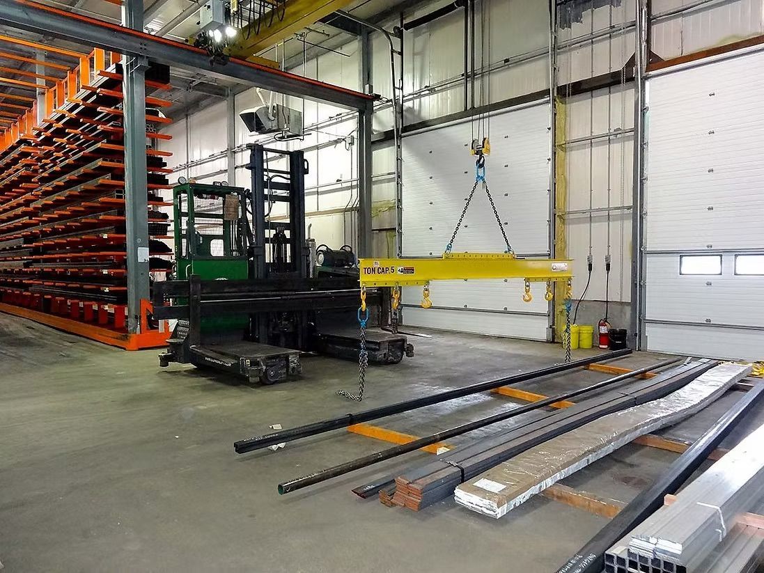 A yellow crane spreader bar hanging above steel beams in an industrial warehouse with storage racks.