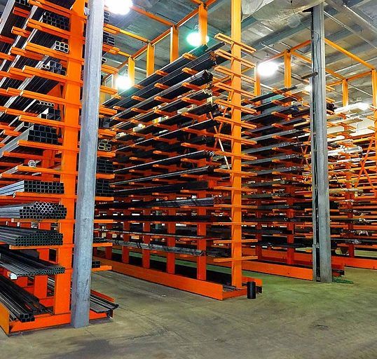 Rows of orange industrial cantilever racks in a warehouse, stocked with various steel beams and metal pipes.