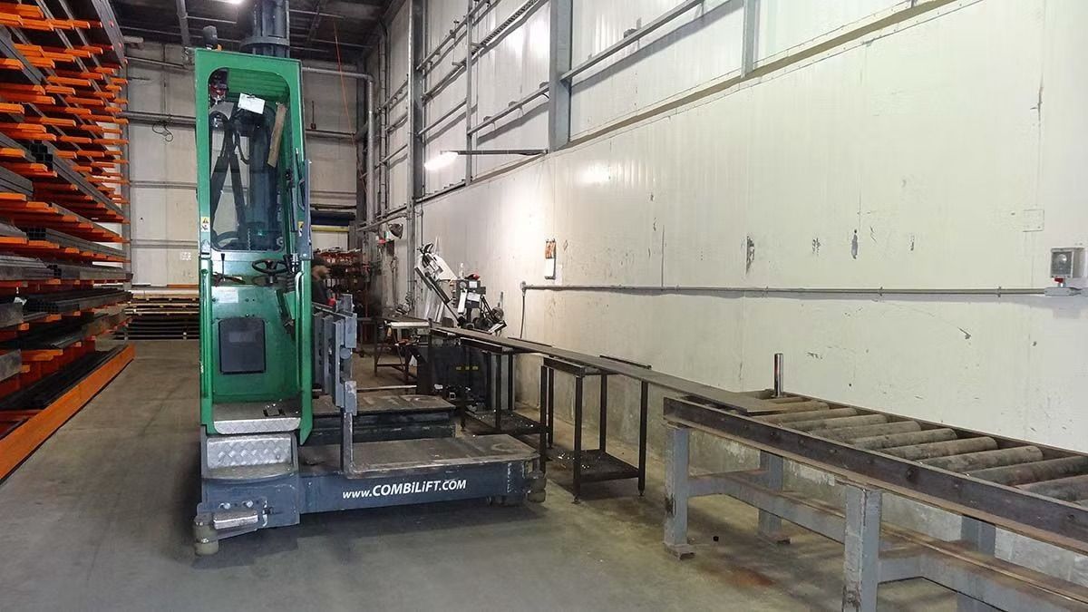 A green industrial forklift positioned next to a metal conveyor table inside a warehouse with tall storage racks.