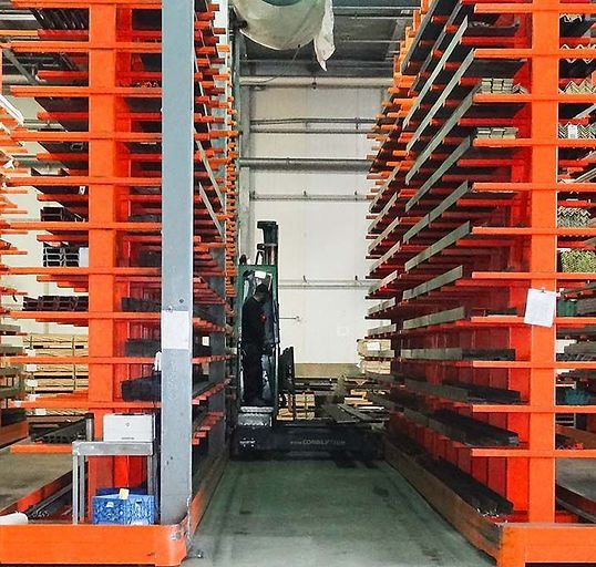 A forklift operates between two tall, orange metal cantilever racking systems filled with stored industrial steel bars.