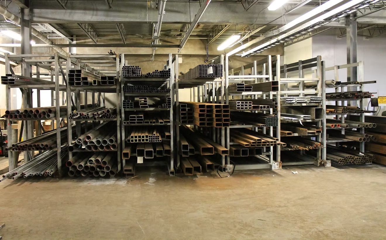 A metal warehouse with rows of heavy industrial steel beams, pipes, and tubing stored on large metal racking units.
