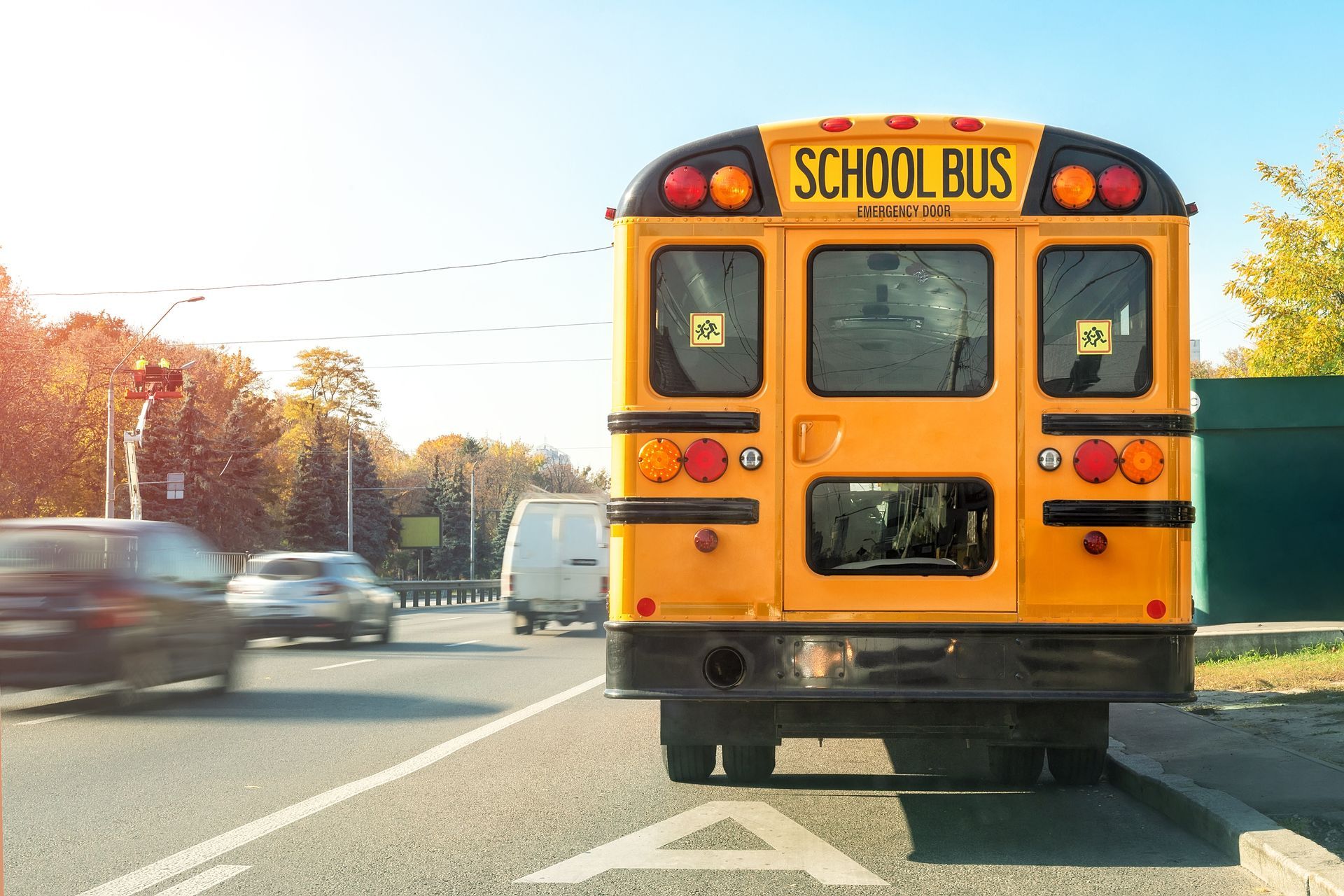 Yellow school bus stopped on a road, back view. Cars in motion. Bright day.