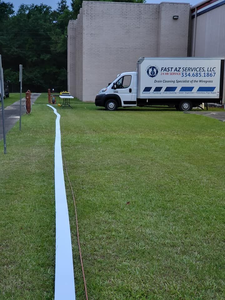 White utility truck parked beside a beige building on a grassy lawn with a long white hose or cable running ahead