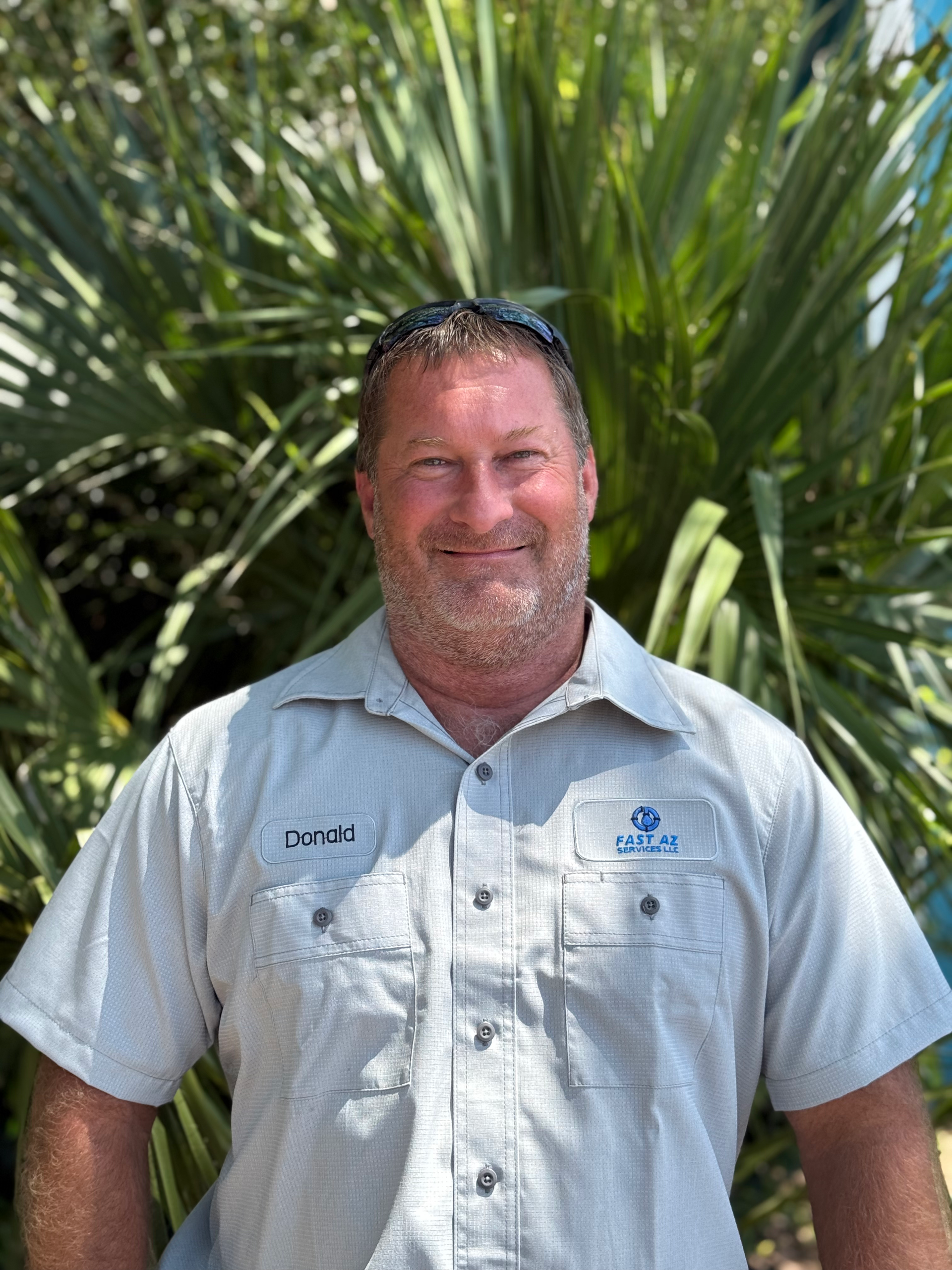 Man in a light gray work shirt smiling outdoors in front of lush green plants