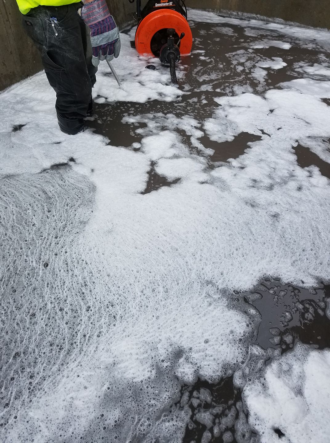 Workers clearing white foam from a wet pavement with a hose and shovel in an outdoor area