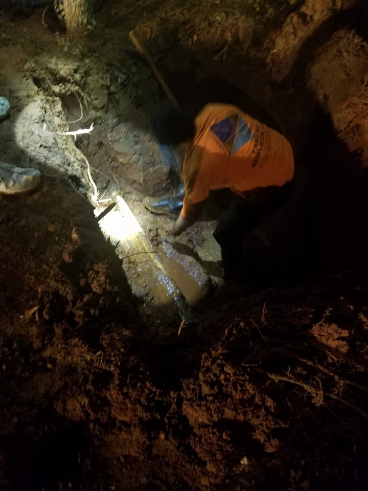 Worker in orange jacket digging in a dark trench lit by a bright work light