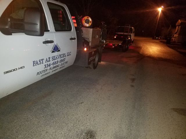 Tow truck parked at night on a dim residential street, with another vehicle ahead and streetlights glowing.