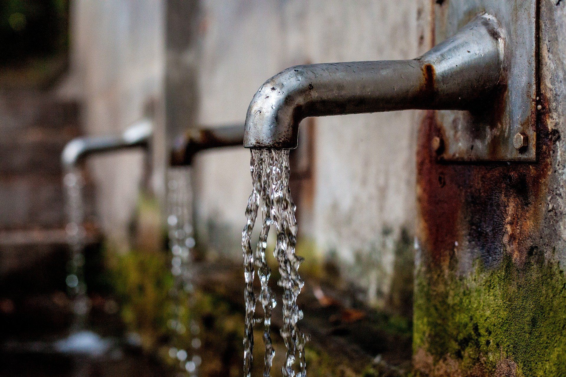 Water flowing from a rusty outdoor metal faucet against a mossy wall.