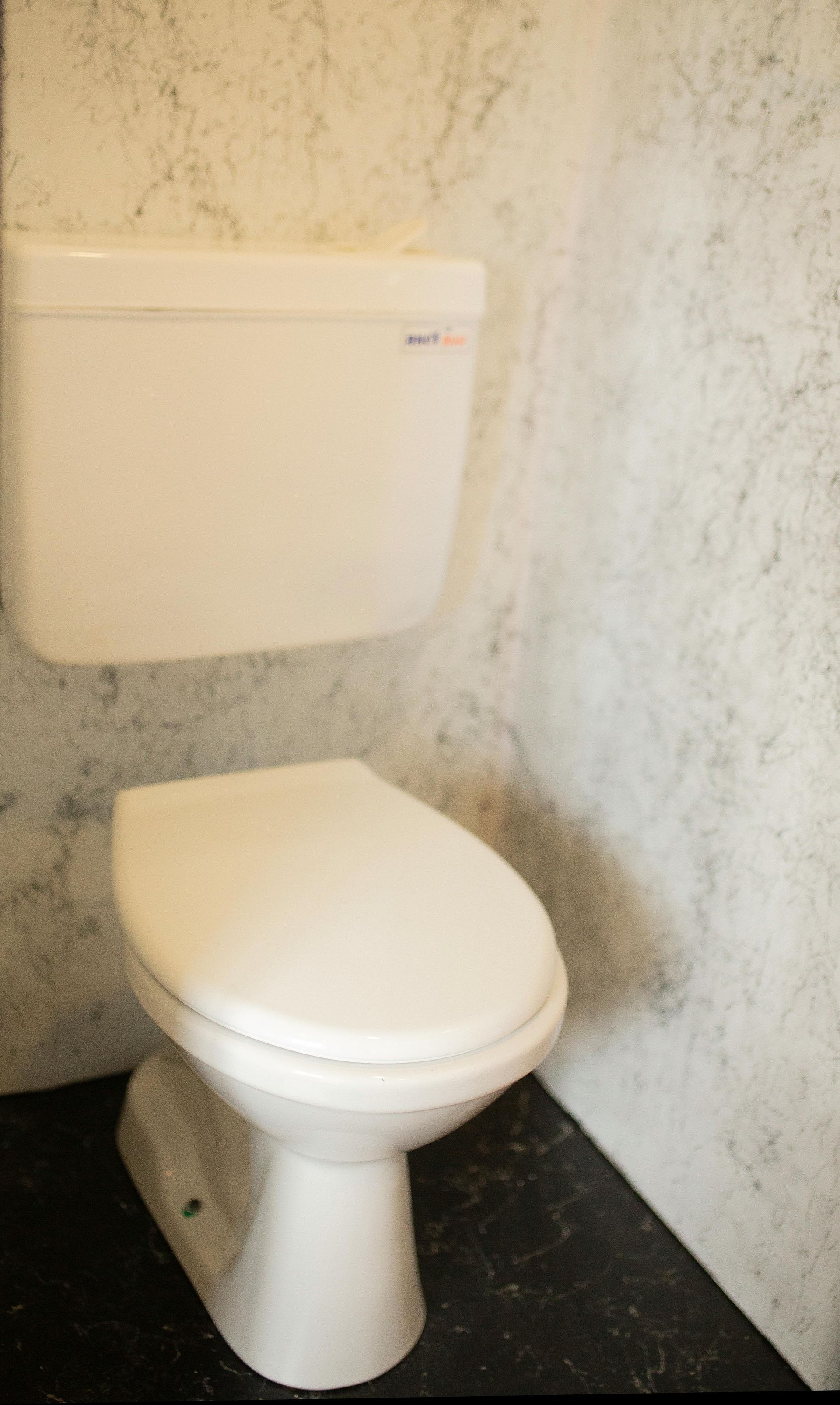 Small white toilet with tank in a plain, textured restroom stall with dark floor.
