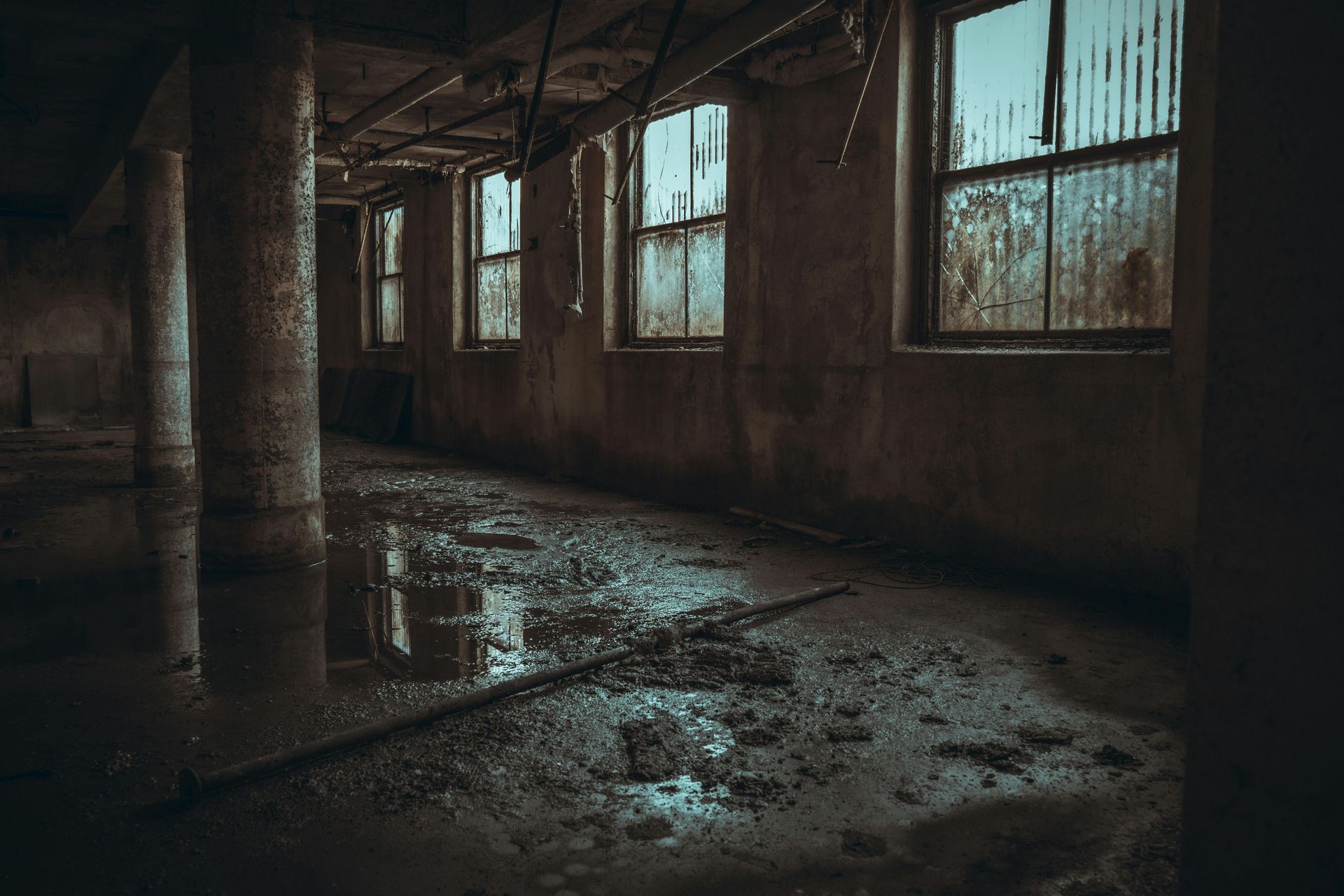 Abandoned industrial room with broken windows, peeling walls, and a wet, debris-strewn floor.