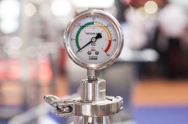 Close-up of a stainless steel pressure gauge with a white dial and colored needles on a blurred industrial background