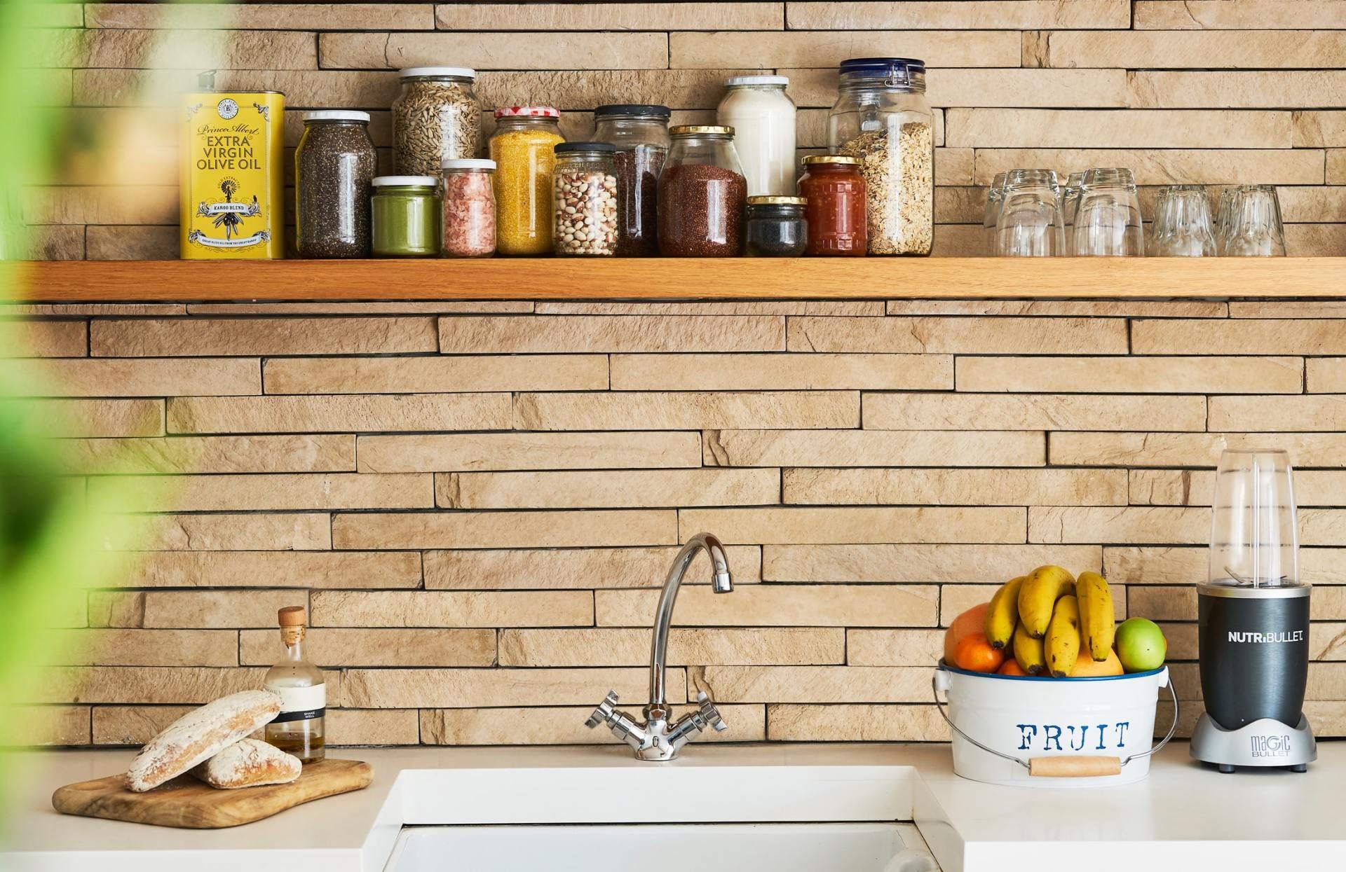 Bright kitchen shelf with jars, fruit bowl, sink, and coffee grinder on a brick backsplash