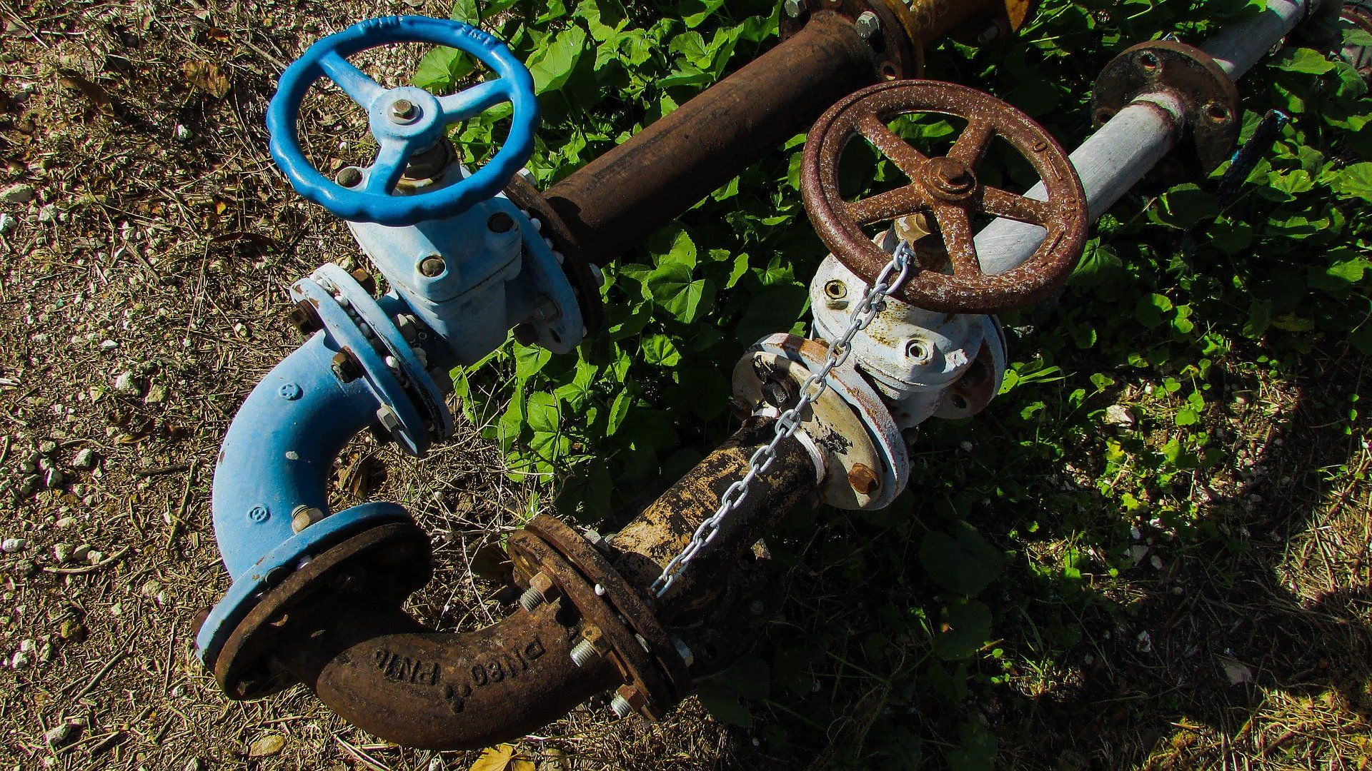 Two rusty pipe valves with blue and brown wheels lying on the ground amid grass and dirt