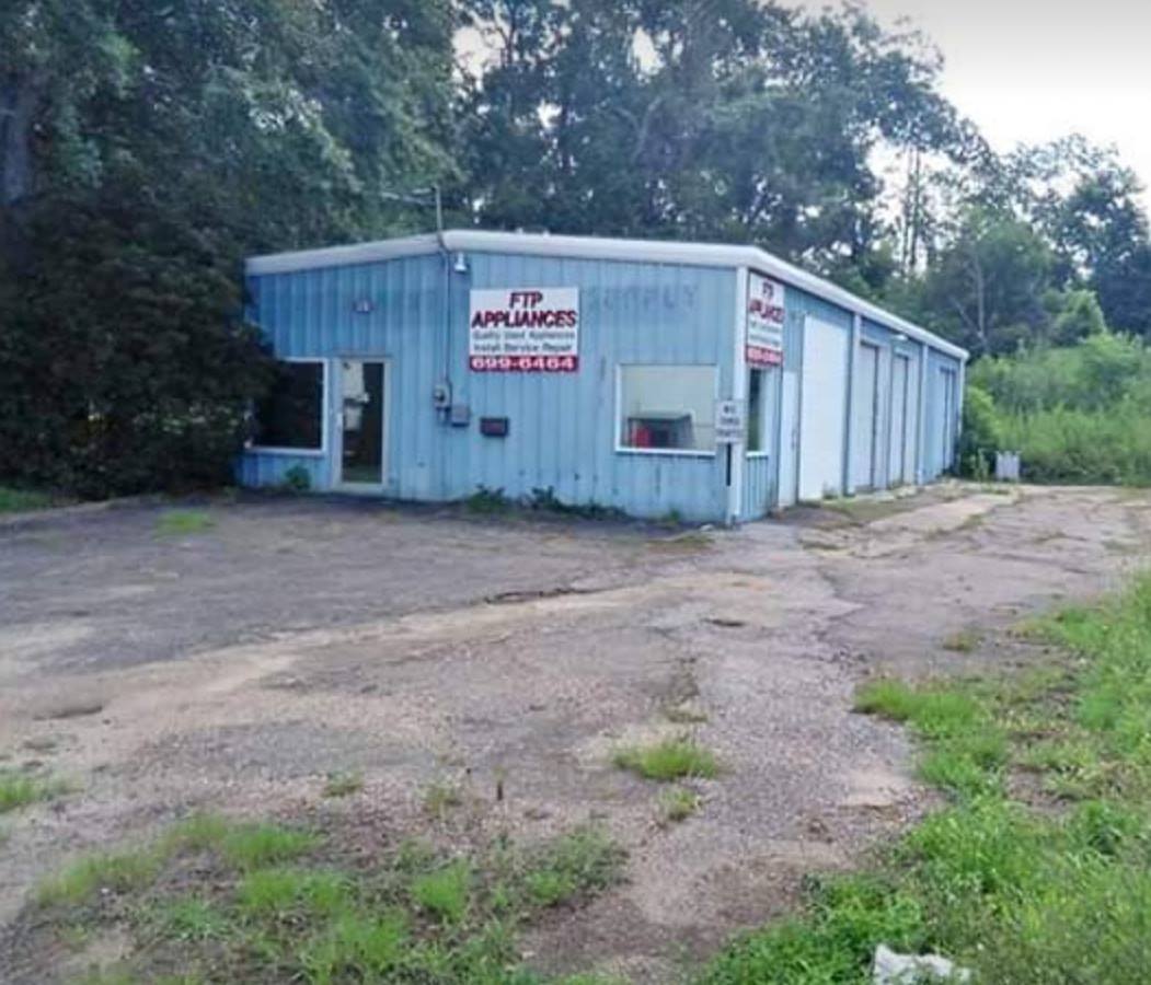 Small light-blue roadside shop building with sign, beside a gravel lot and trees.